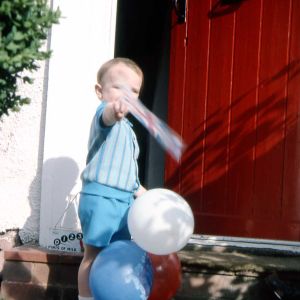 Dominic as child, waving a flag with some ballons in front of a red door.